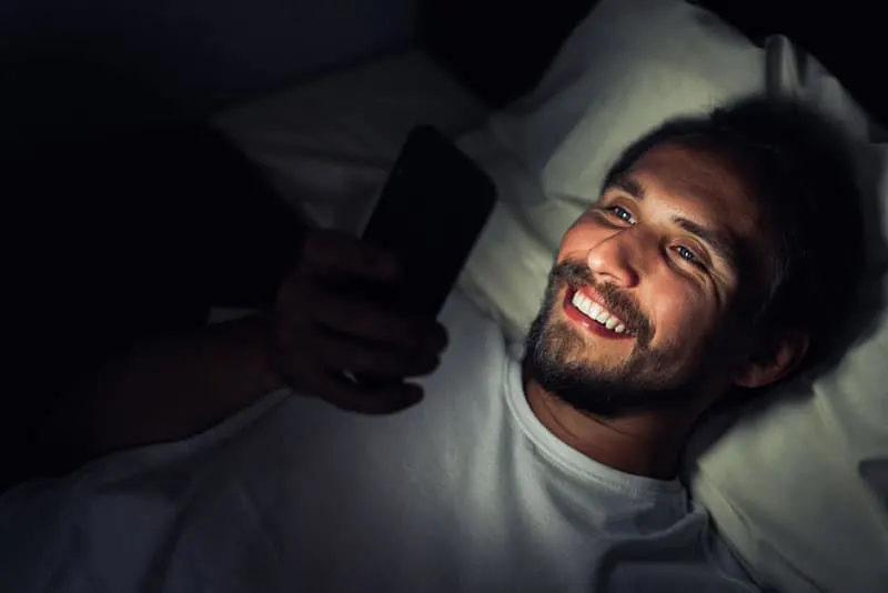 smiling man looking at his phone while lying on bed