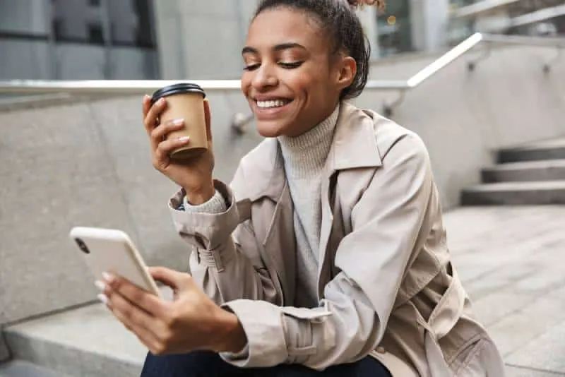 smiling woman holding typing on her phone while holding a cup