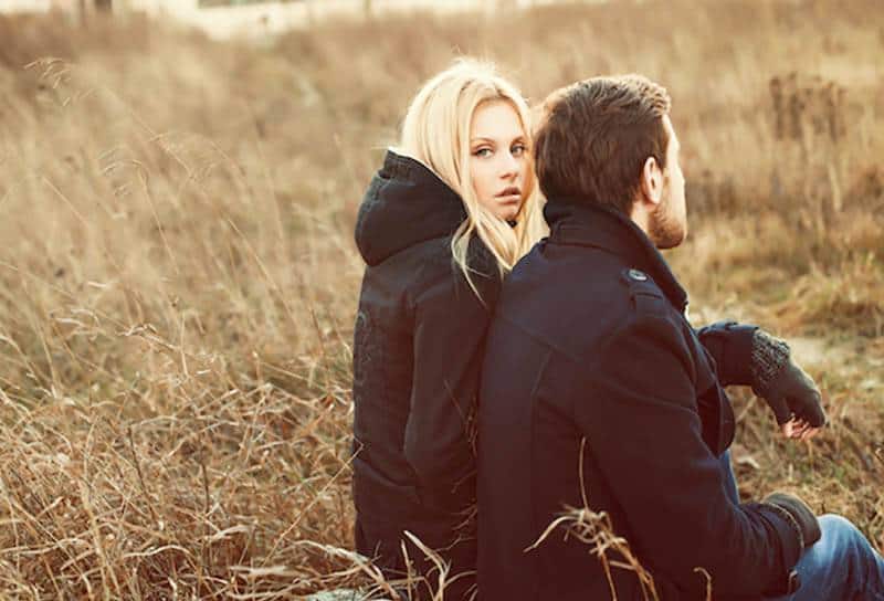 thoughtful blonde woman sitting next to man wearing black jacket and gloves outside