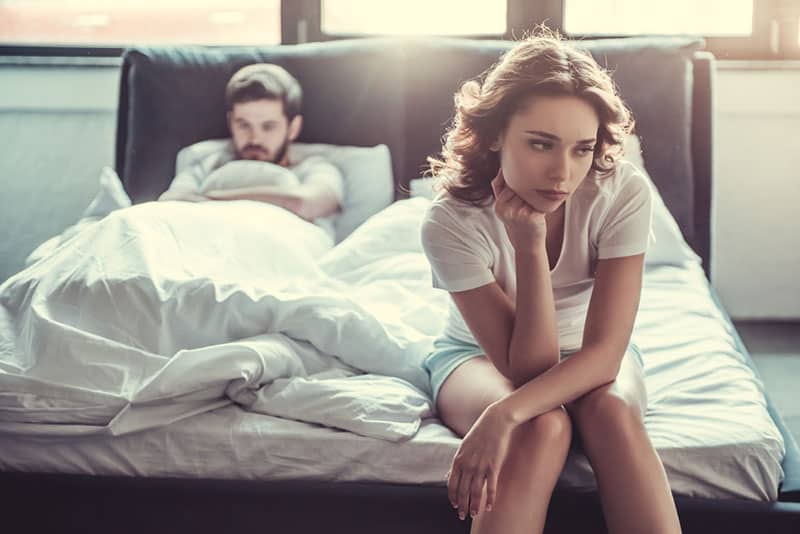 upset woman sitting on bed while man lying on bed