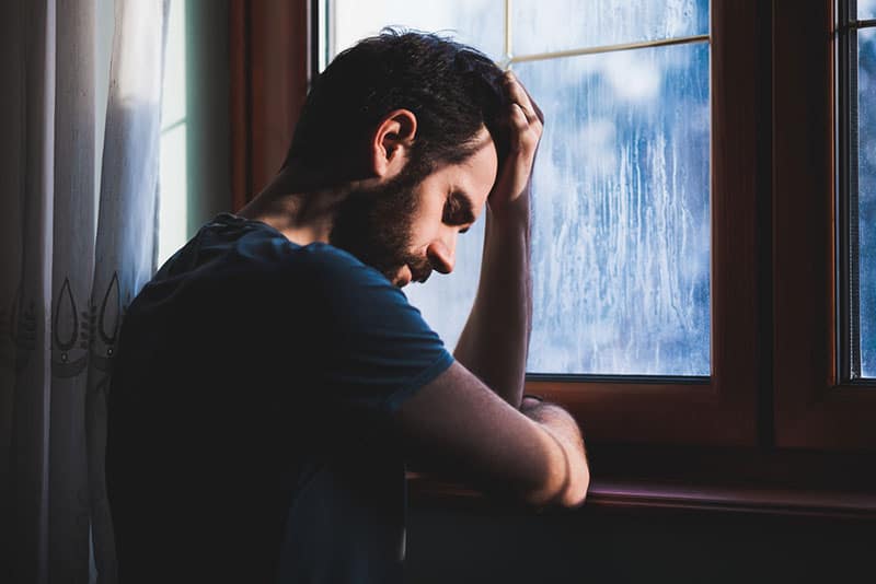depressed man standing in front of window at home