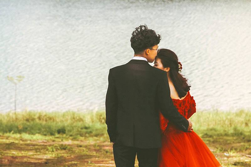 man in black suit kisses woman in red dress beside water