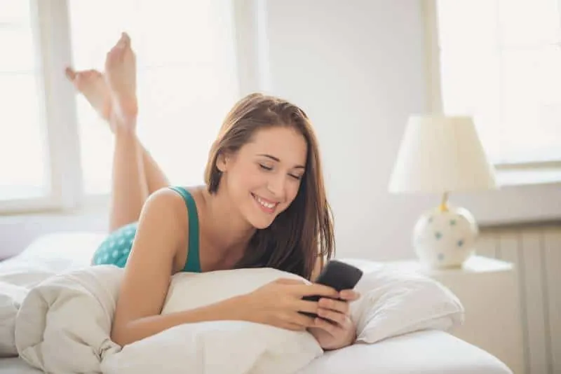 smiling brunette typing on her phone while lying on bed