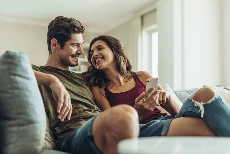 smiling couple sitting on the couch and looking at phone