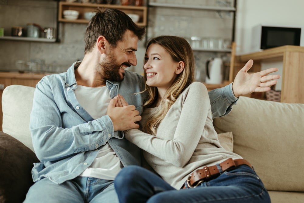 a smiling couple sitting on the couch hugging and talking