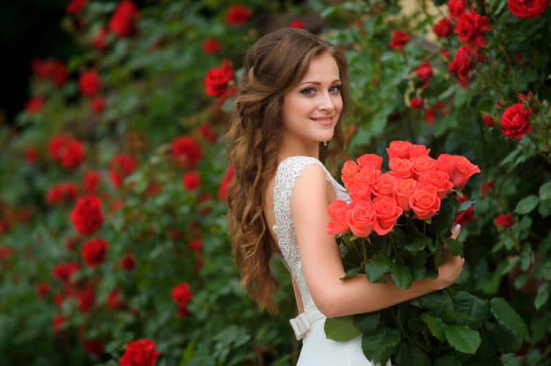 rapariga bonita com cabelo comprido e vestido branco segurando um balde de rosas vermelhas