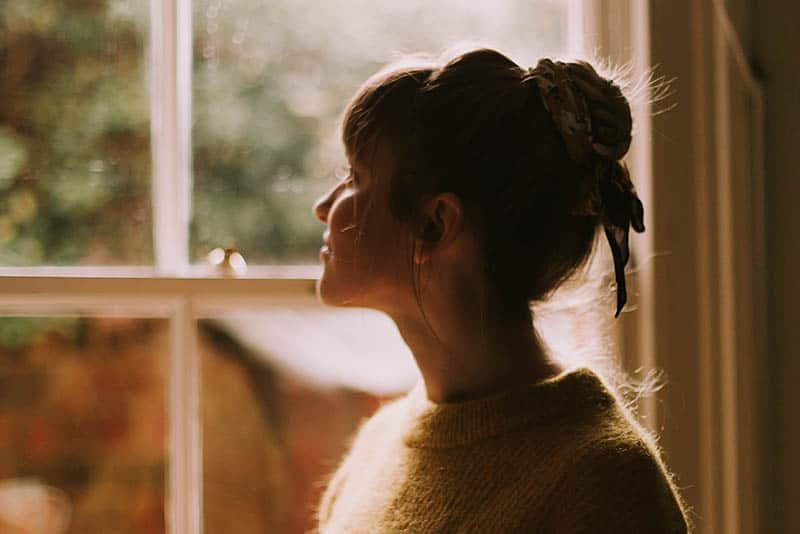 vista laterale di una donna con i capelli legati in piedi accanto alla finestra e che guarda fuori