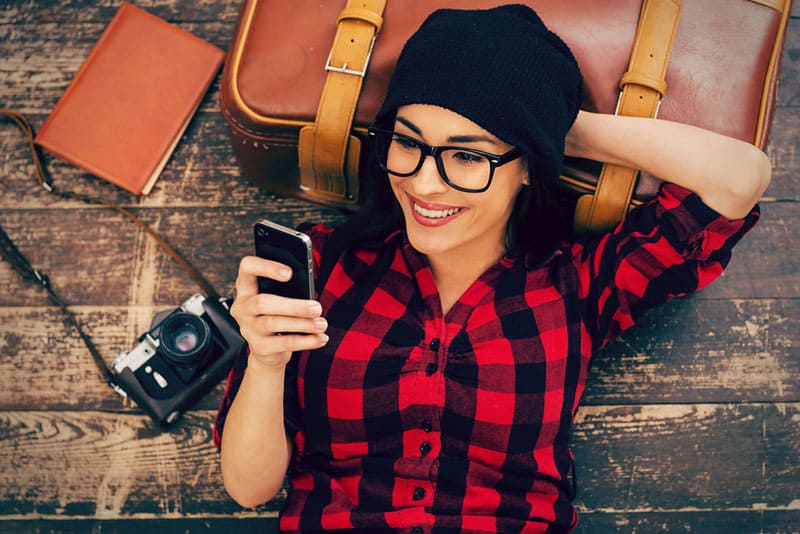 mujer sonriente con camisa de cuadros y apoyada en el suelo de madera y tecleando en su teléfono