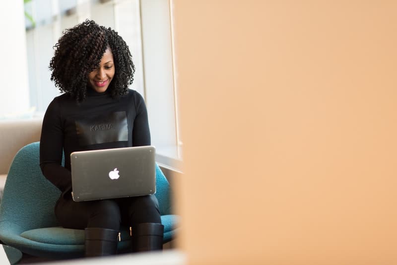 a smiling black woman sits and uses a laptop