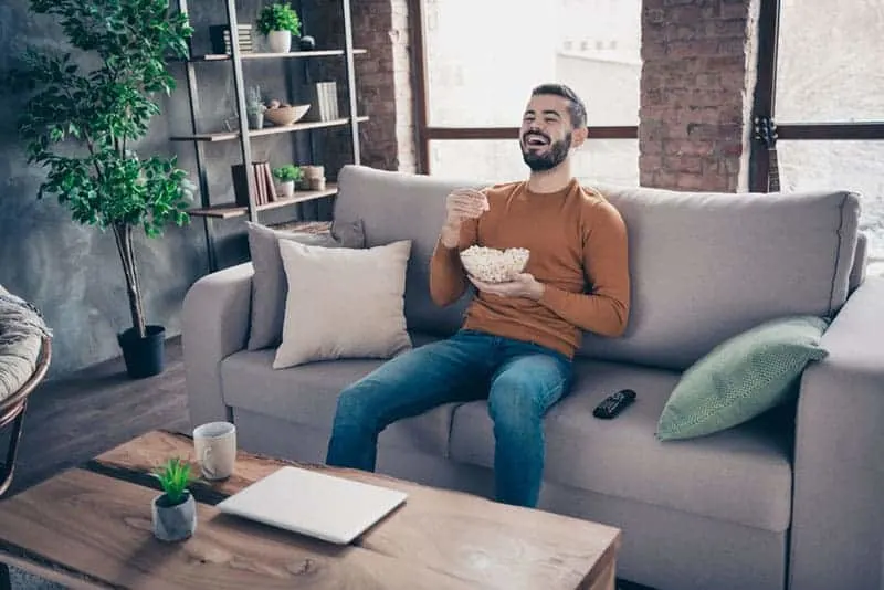 man eating popcorn and laughing while watching a TV