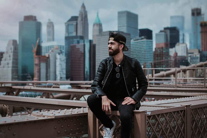  homme en veste de cuir noir debout au sommet d'un pont avec des bâtiments de la ville derrière lui