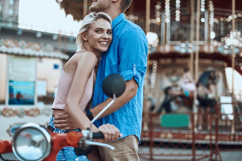 uomo con camicia blu che abbraccia una giovane donna bionda e sorridente all'esterno