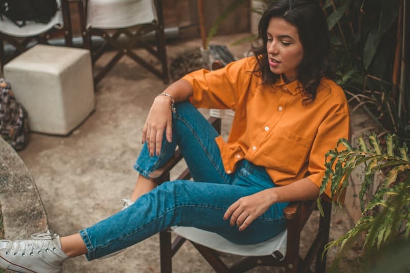 outside on a wicker chair sits a girl in an orange shirt and levis pants