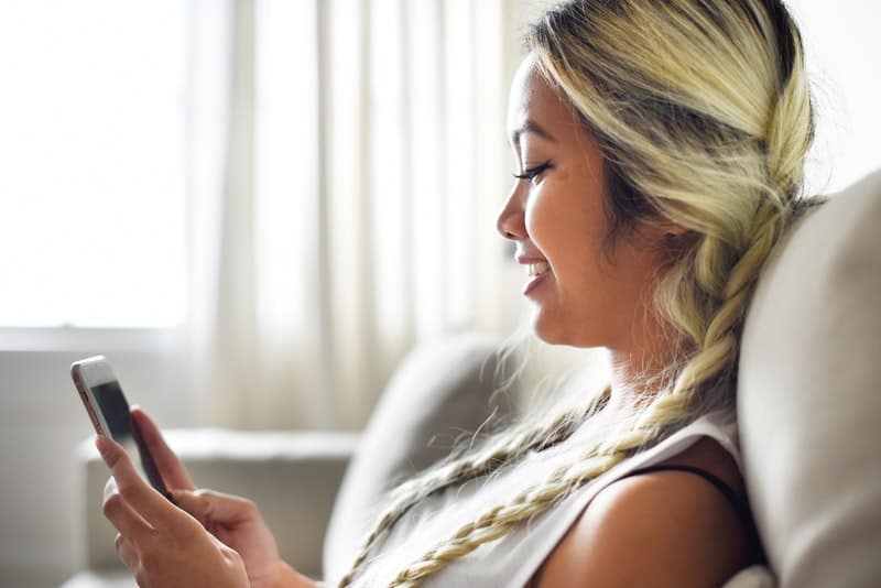 donna asiatica sorridente che guarda il suo telefono mentre è seduta sul divano