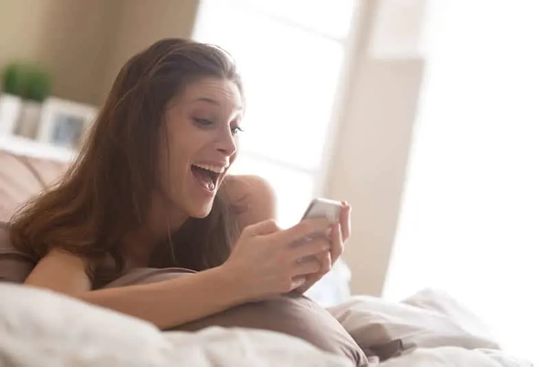smiling woman lying on bed and typing on her phone