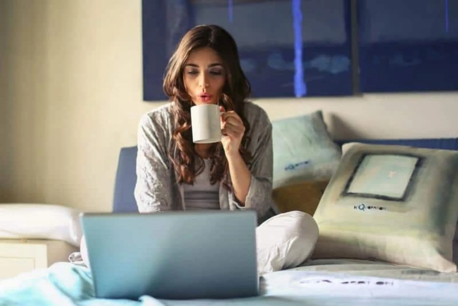 woman sitting in bed drinking coffee and typing on laptop