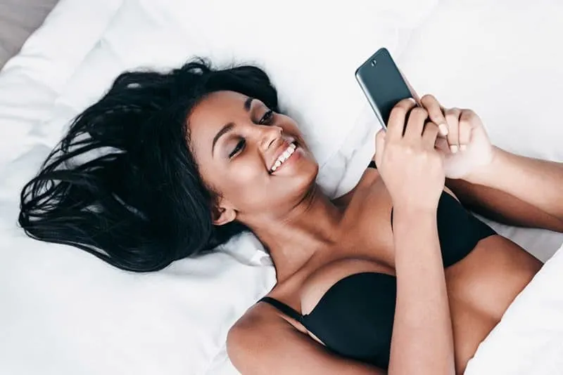 young smiling woman lying on bed and typing on smartphone