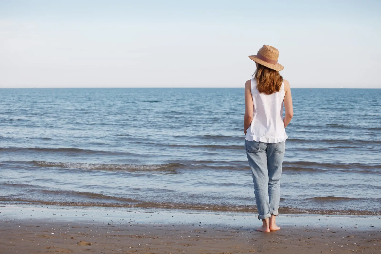 a barefoot woman with a hat standing on the beach by the sea