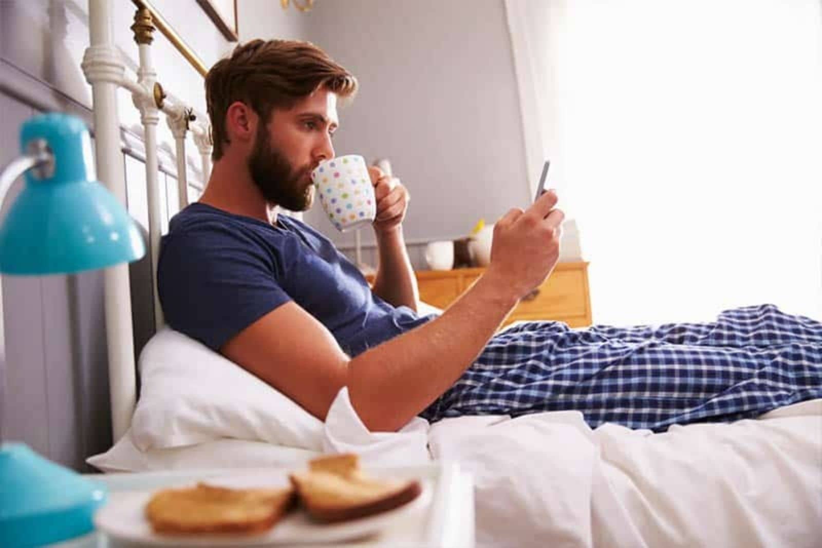 a man sitting in bed drinking coffee and pressing a phone