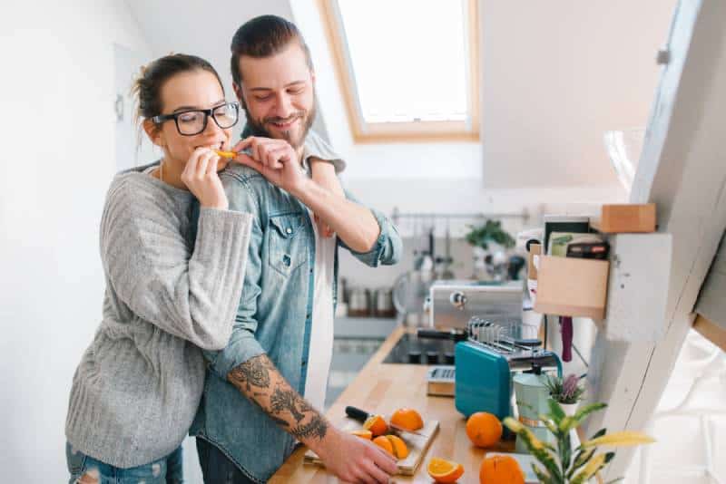 man giving piece of orange to his girlfriend at the kitchen