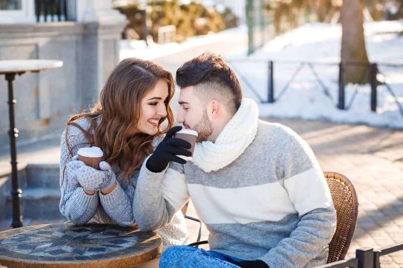 smiling couple wearing sweatshirt and gloves while holding cup of coffee at street cafe