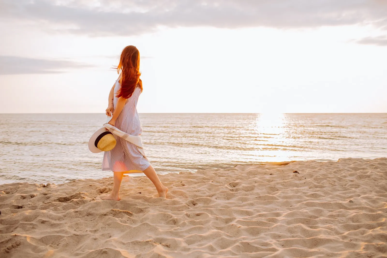 woman in a dress holding straw hat and walking alone on empty sand beach