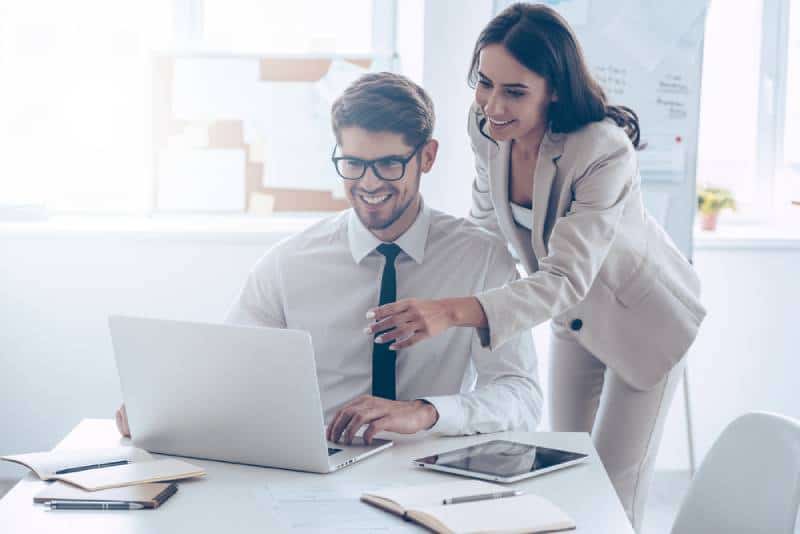 mujer señalando a la computadora portátil con una sonrisa con su compañero de trabajo mientras está de pie en la oficina