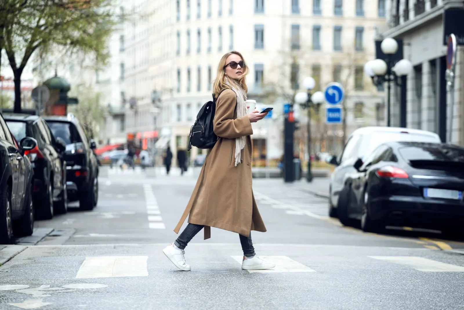 a blonde in a coat with sunglasses crosses the street