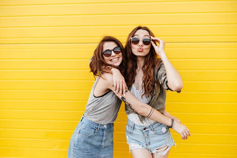 happy female friends wearing sunglasses and posing for camera with yellow background