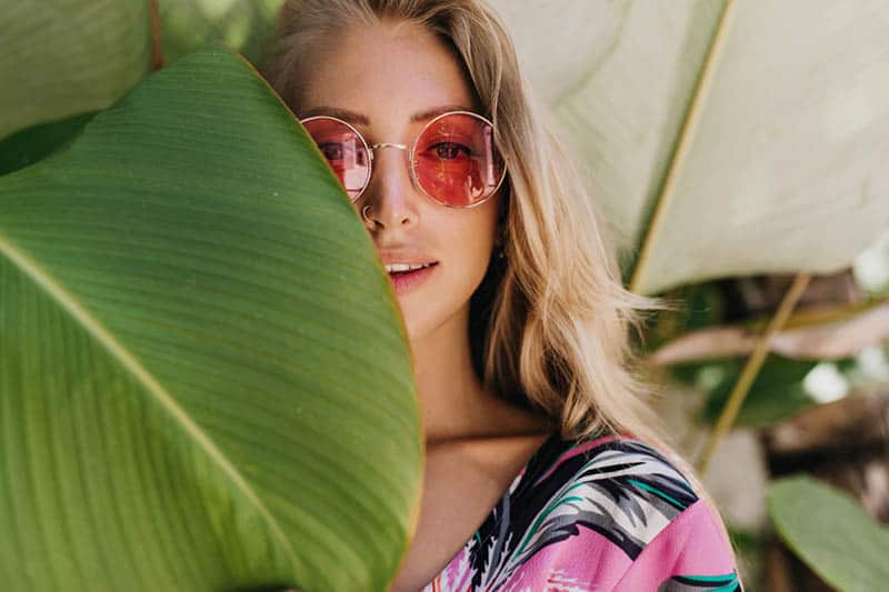 retrato de mujer con gafas de sol rojas de pie junto a una hoja