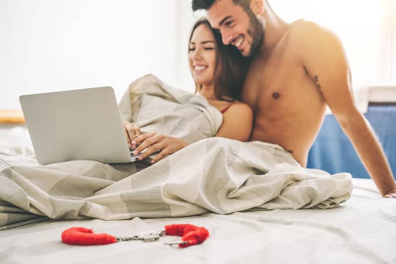 Smiling naked couple under blanket looking at laptop