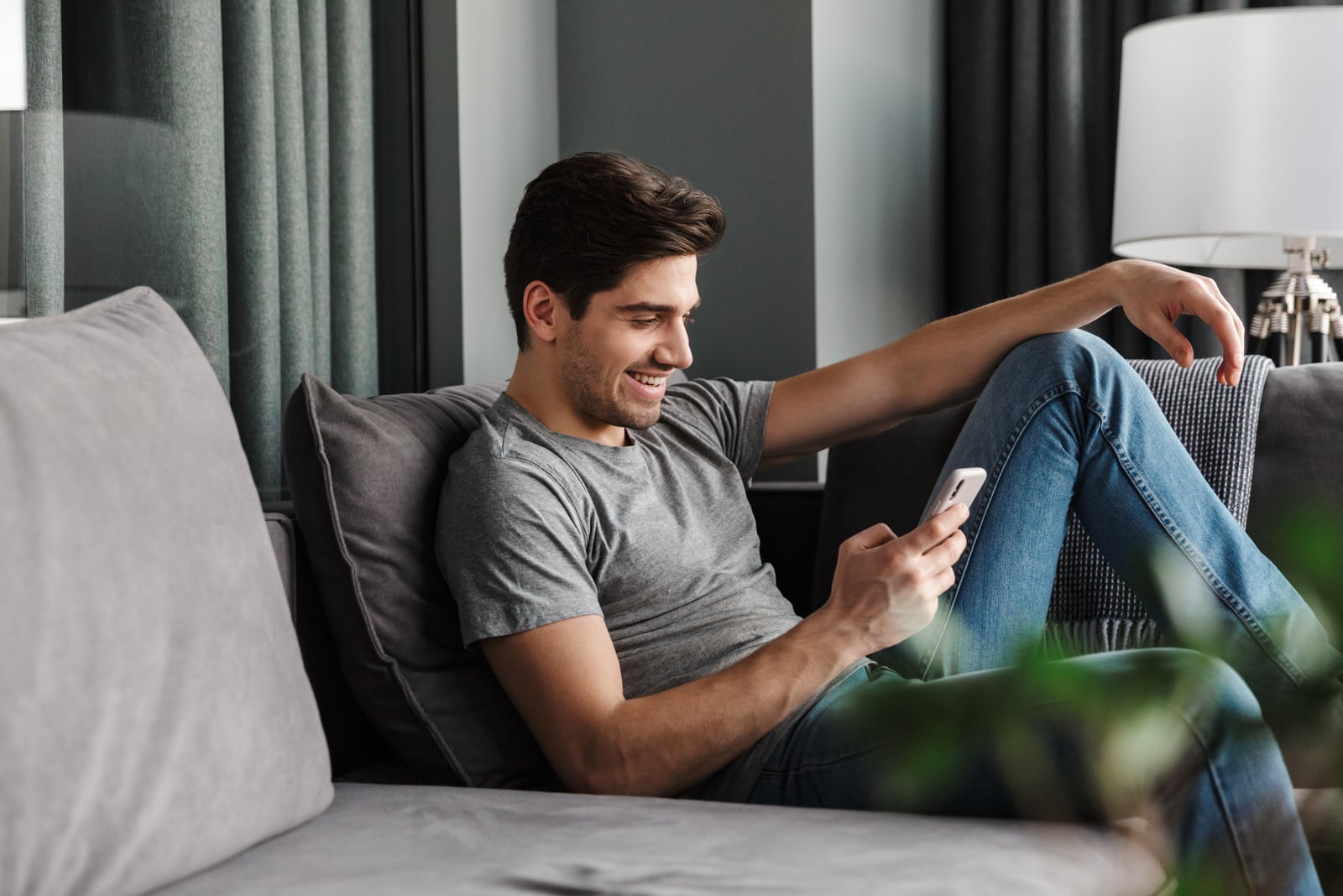 an attractive smiling man sits on a sofa and uses a smartphone