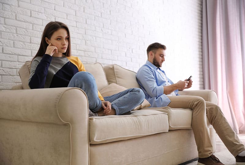 sad woman sitting on the couch while man typing on phone