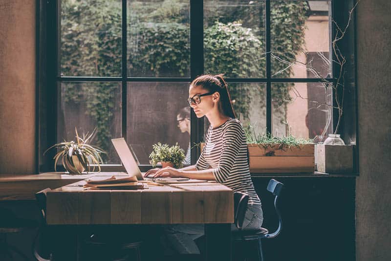 woman sitting next to window and typing on her laptop