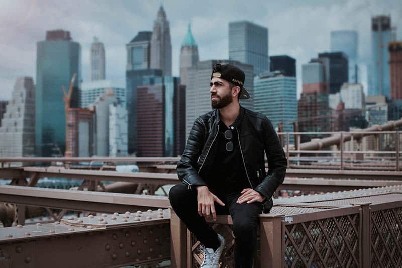 man in black leather jacket standing on top of bridge with city buildings behind him