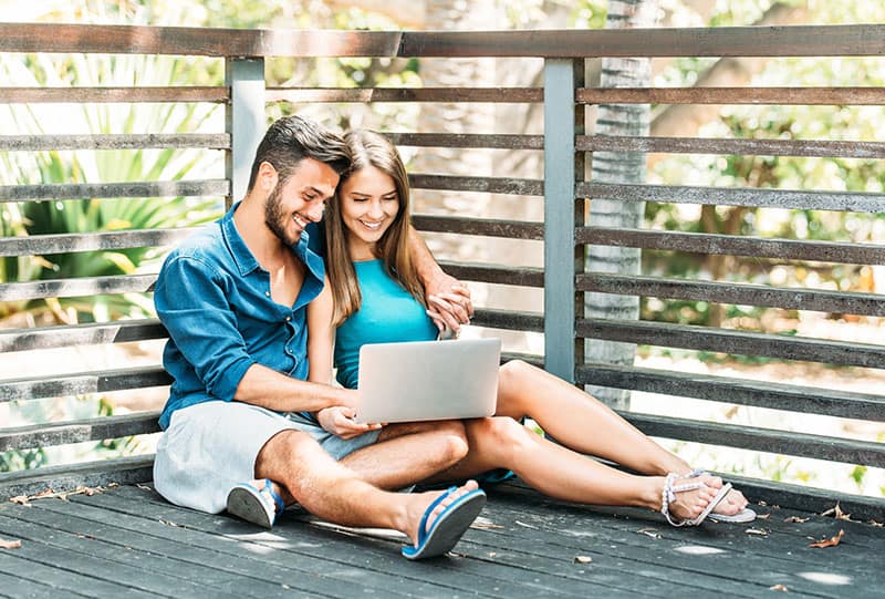 Young happy couple shopping online on their laptop sitting on wooden floor outside
