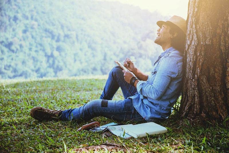 smiling man leaning on tree and writing on paper