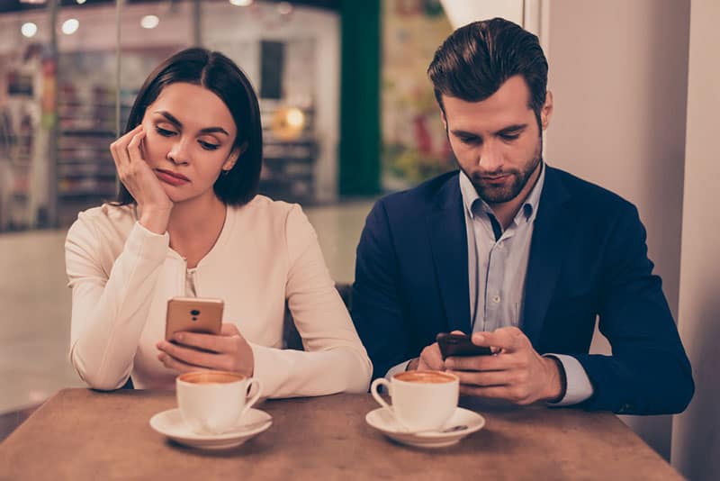 Pareja aburrida sentada en un café sosteniendo teléfonos