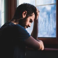 depressed man leaning hand on window at home