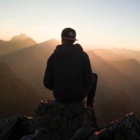 vista traseira de um homem sentado no cimo de uma colina e de frente para as montanhas