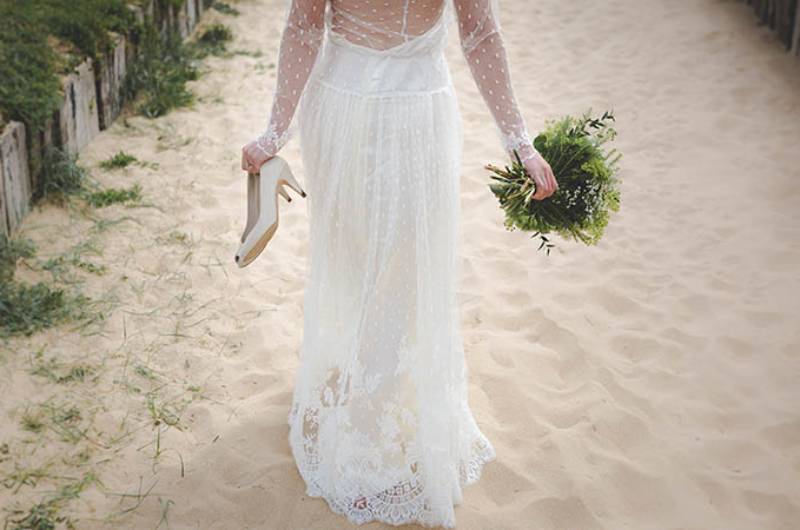 donna sposa che cammina sulla sabbia con fiori e tacchi in mano