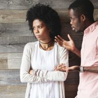 Young displeased black male gesturing in indignation, arguing with his stylish girlfriend, who is standing against wooden wall with folded arms, looking away with offended expression on her face