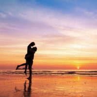 Coppia che si bacia sulla spiaggia con un bellissimo tramonto sullo sfondo, uomo che solleva la donna