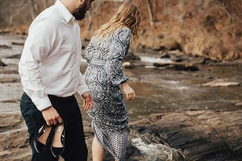man holds woman heels while she walking on wet rocks