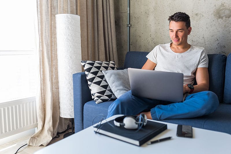 young attractive man sitting on sofa at home working on laptop online