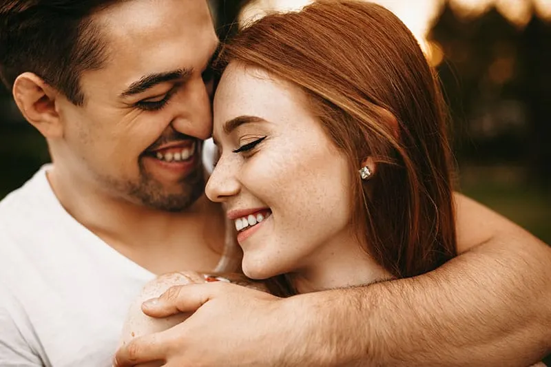Close up portrait of a amazing young female with red hair and freckles smiling with closed eyes while being embraced from back