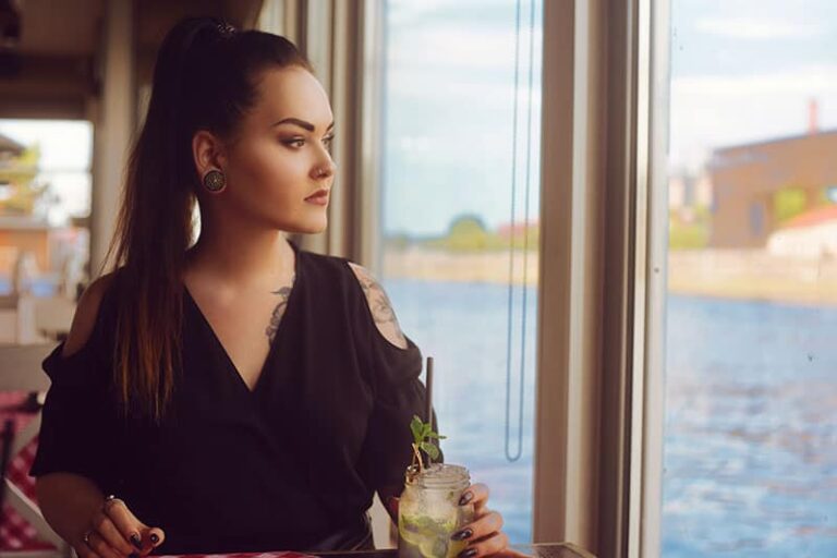 La chica en un restaurante en el barco cerca del río bebe mojito. Morena con tatuaje mira por la ventana y bebe mojito en una jarra de cristal. La chica con túneles en las orejas.