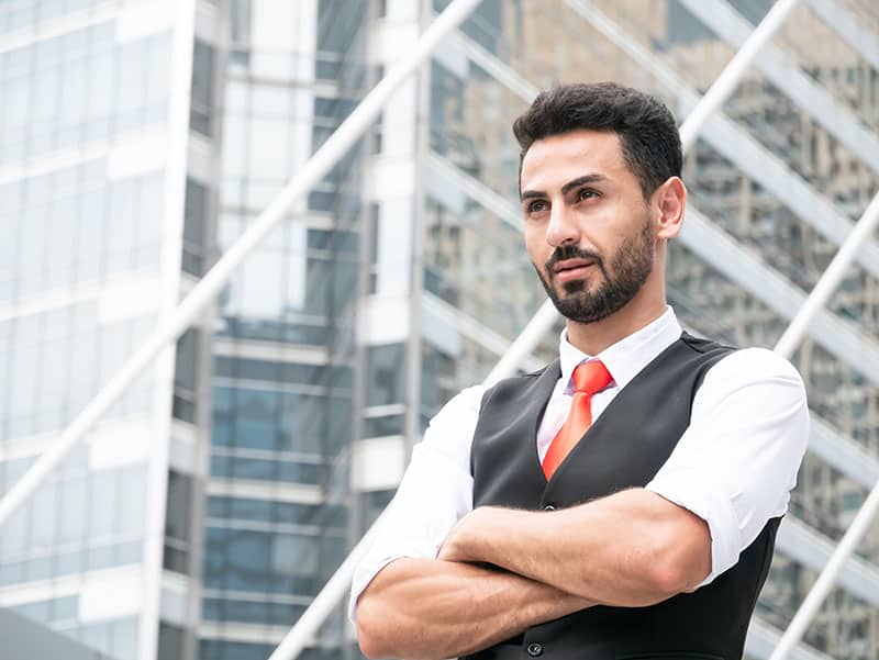 Ritratto di uomo d'affari arabo sicuro di sé in piedi con le braccia incrociate all'aperto davanti a un ufficio moderno.