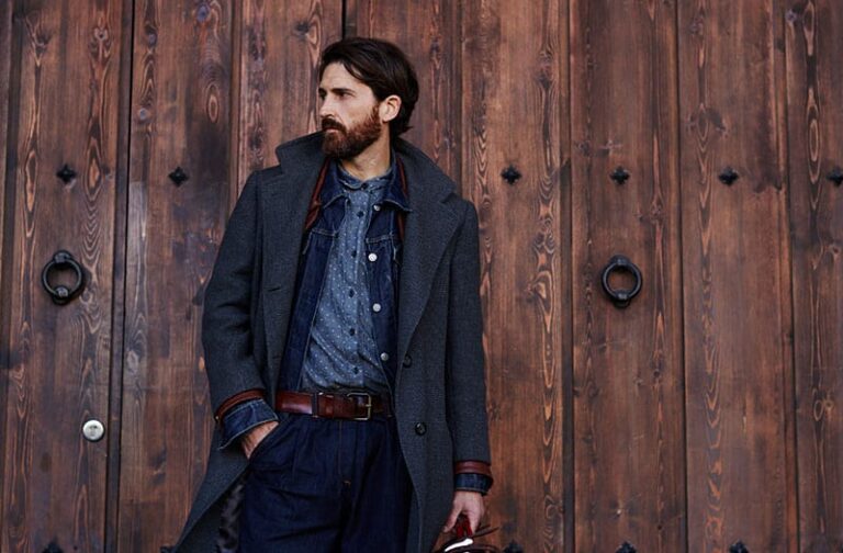 Handsome confident mature man with beard stands on wooden brown background with copy space for your text message or advertising, fashionable rich male dressed in expensive clothes posing outdoors