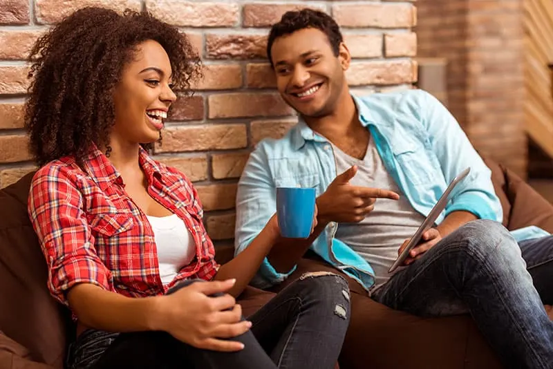 Attractive Afro-American couple using tablet, holding a cup and laughing while sitting on beanbag chairs against brick wall